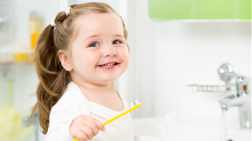 Happy young girl holding a yellow toothbrush while brushing her teeth at the bathroom sink.